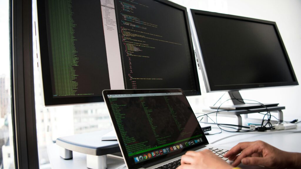 A developer writes code on a laptop in front of multiple monitors in an office setting.